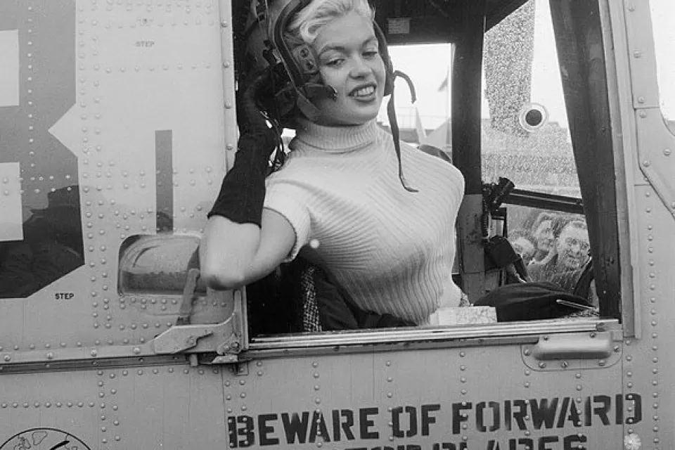 Black and white photo of actress Jayne Mansfield leaning out of the window of a military plane wearing flight helmet for promotional photo